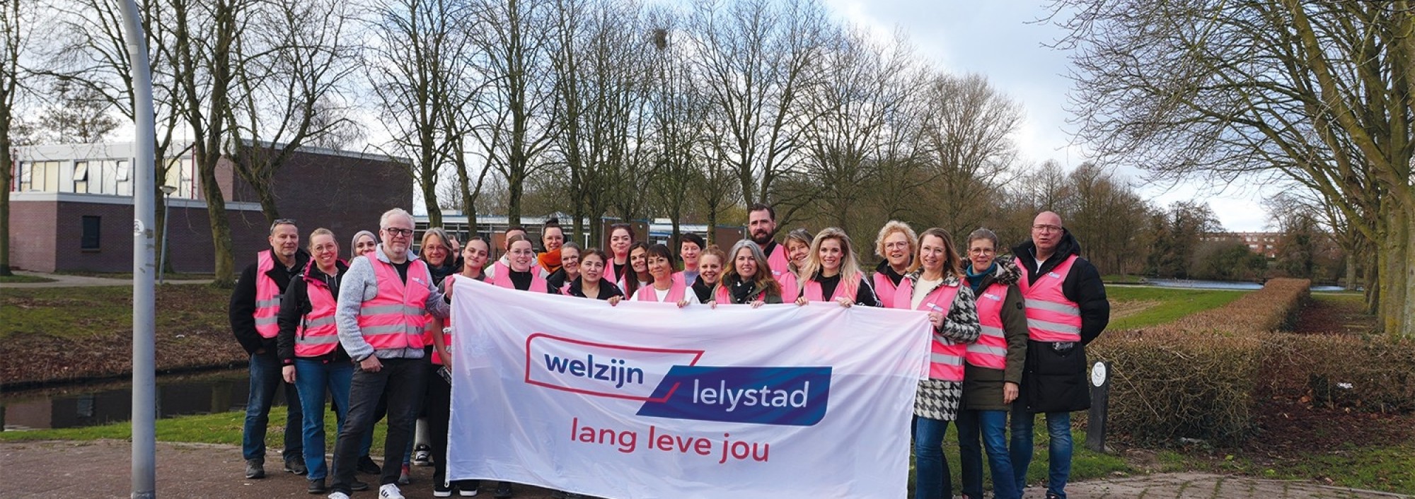 Mannen en vrouwen in roze hesjes houden vlag met logo Welzijn Lelystad vast.