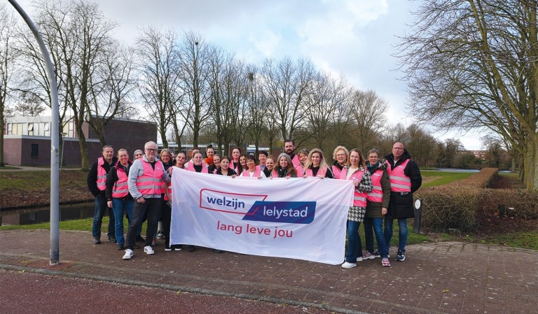 Mannen en vrouwen in roze hesjes houden vlag met logo Welzijn Lelystad vast.