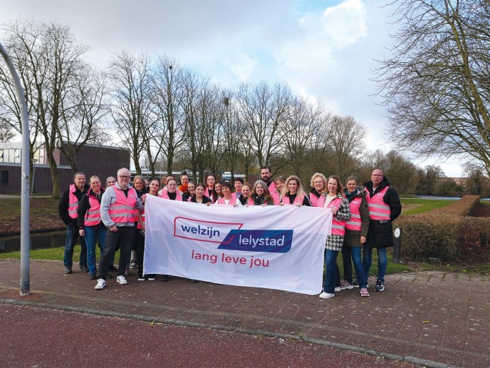 Groep mensen in roze hesjes houdt vlag met logo Welzijn Lelystad vast.